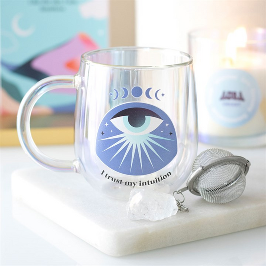 A clear mug with an all-seeing eye design features crescent moons, stars, and the phrase “I trust my intuition.” Nearby are a crystal tea infuser, a white crystal, and a lit candle on a white surface.