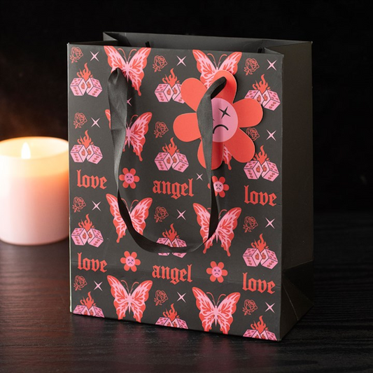 A pop-punk gift bag with red and pink designs—roses, flames, ‘love’ and ‘angel’ text, a cybersigil butterfly, and a tag with a sad face. A lit white candle stands behind it on a dark surface.
