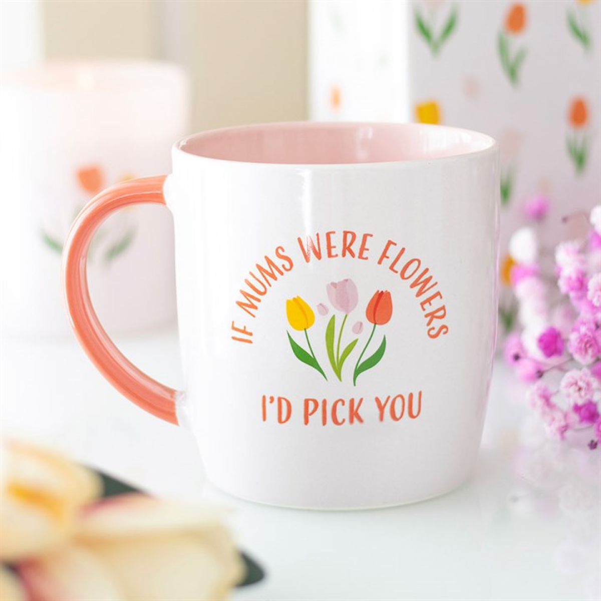 A ceramic Mother’s Day mug with a pink handle and interior displays three pastel tulips and the phrase “If mums were flowers I’d pick you.” An ideal gift for mum, with floral details and blurry shapes in the background.