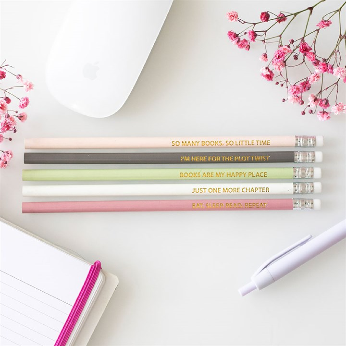 Five pastel pencils adorned with literary quotes rest beside a white mouse, pink flowers, an open notebook with a pink elastic, and a white pen—a lovely present for book lovers on a white surface.