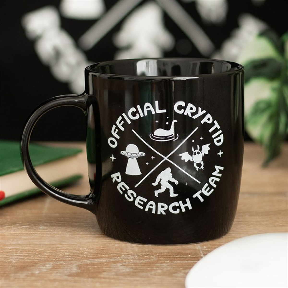A black ceramic mug rests on a wooden table, displaying white text that reads "Official Cryptid Research Team" alongside icons of a UFO, Bigfoot, Mothman, and Nessie. A plant and a book are blurred in the background.