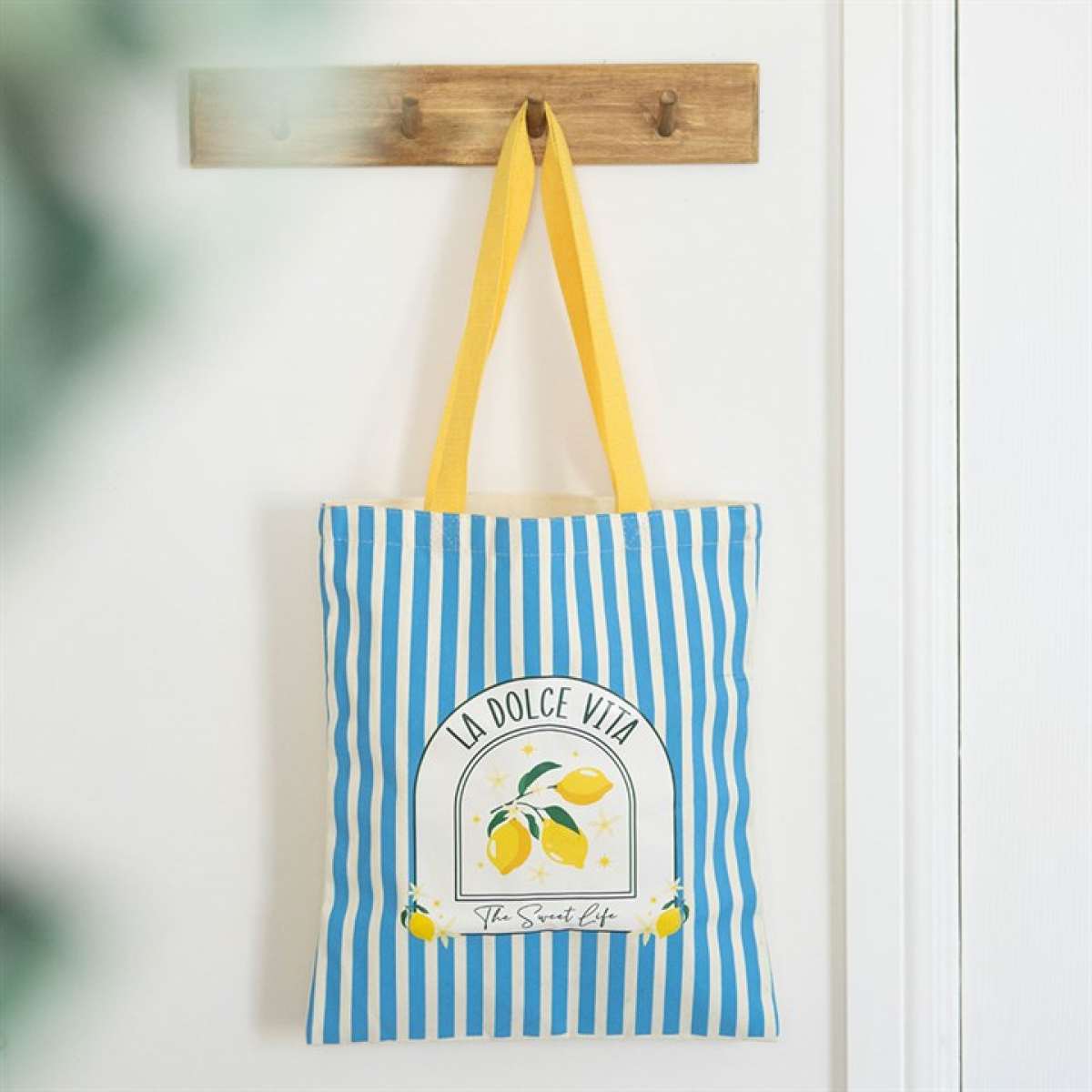 A nautical tote bag with blue and white vertical stripes hangs from a wooden hook. It has yellow handles and displays lemons with the words "La Dolce Vita" and "The Sweet Life" on the front.