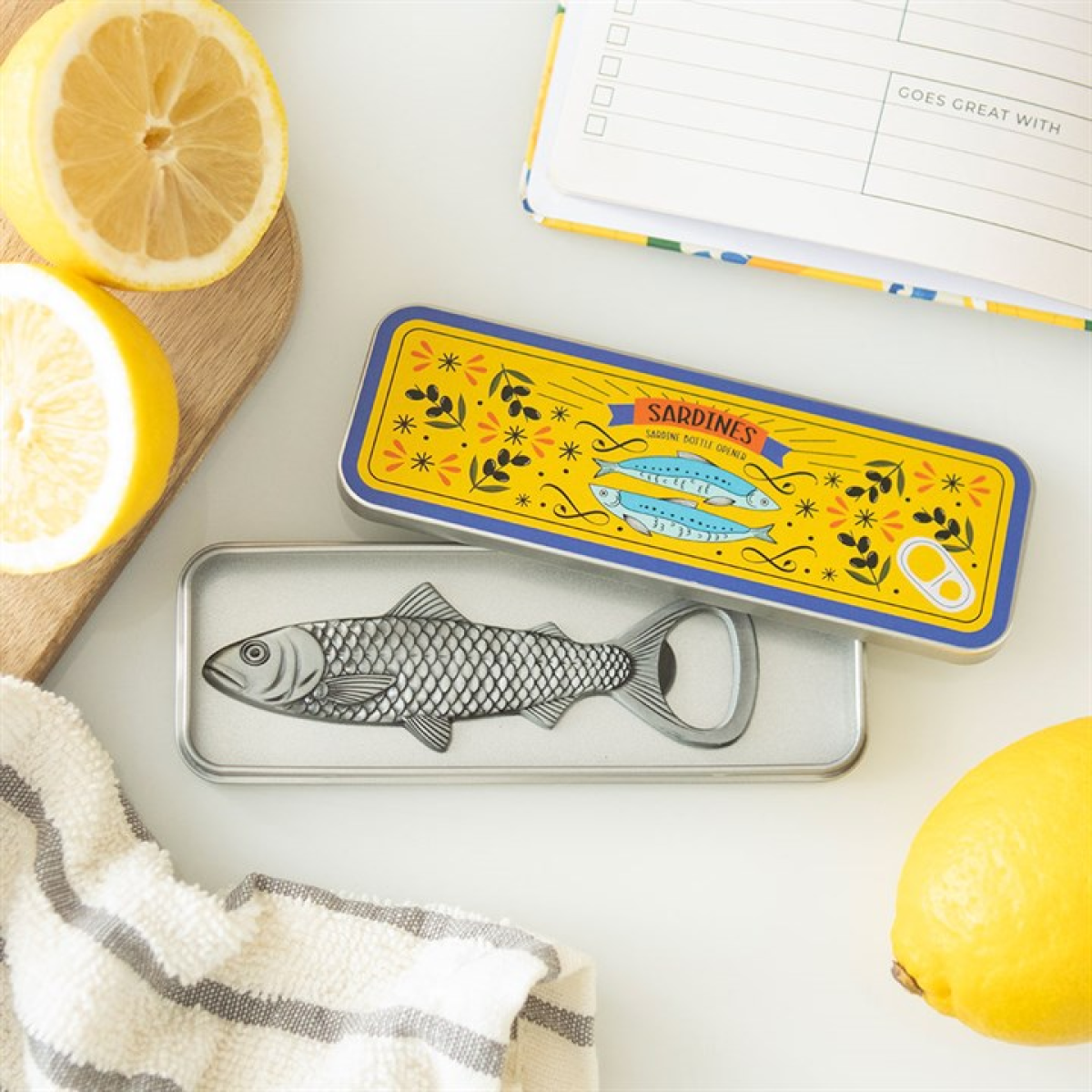 A nautical barware accent, this sardine-shaped novelty bottle opener sits in an open sardine tin adorned with flowers and fish. Nearby on the white surface lie lemons, a notebook, and a striped towel.