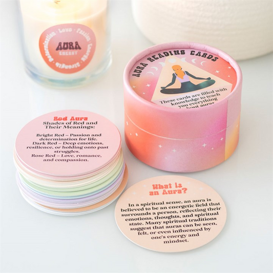 A pastel-coloured box labelled Aura Reading Cards rests on a white surface, its round cards fanned out for spiritual exploration. One card explains auras; another details red aura meanings. A candle labelled Aura Energy glows behind.