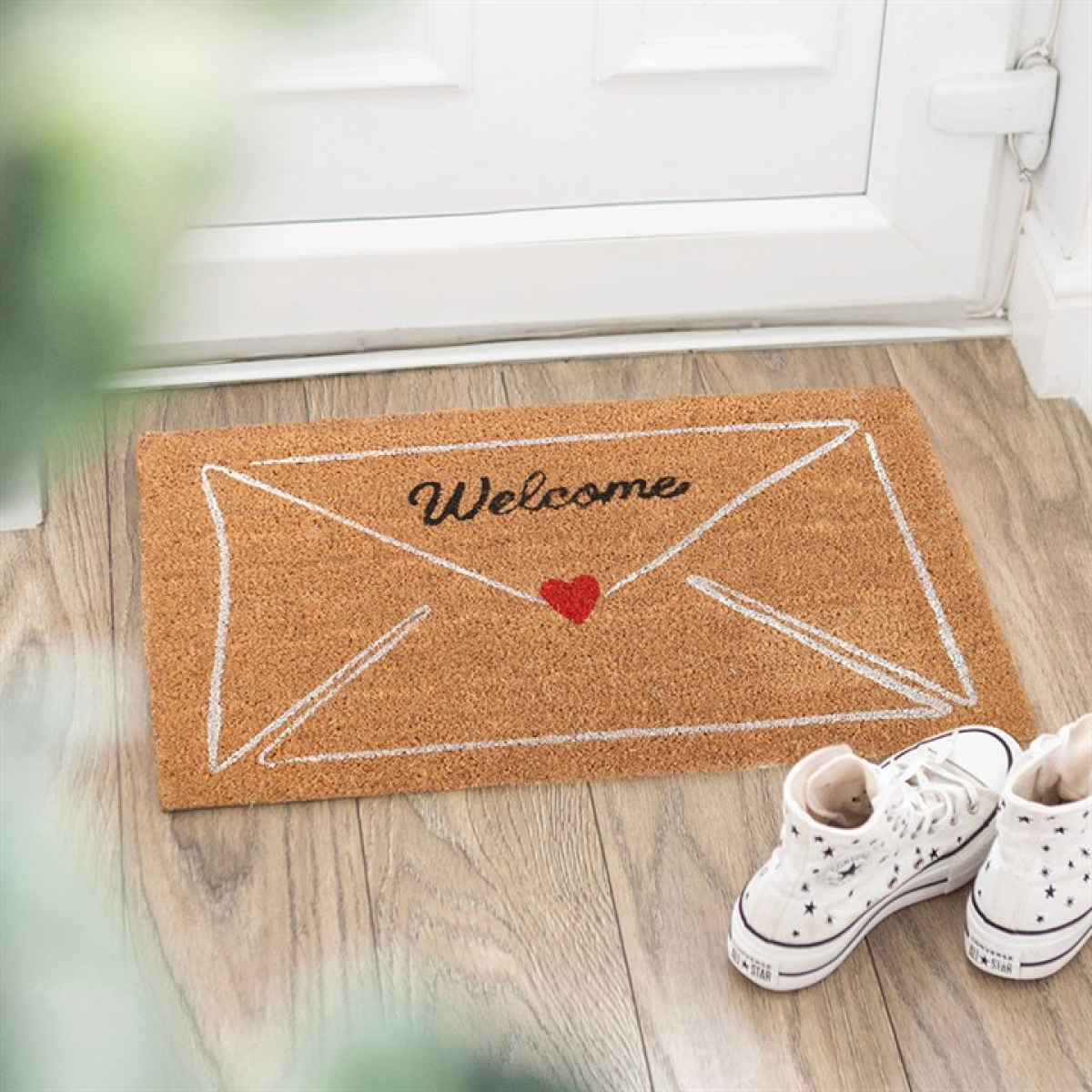 A love letter envelope doormat with “Welcome” and a red heart rests on wooden flooring beside a white door, ideal for Valentine’s Day. Close by, white trainers with star patterns lend a playful accent.