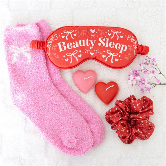 A flat lay featuring a red “Beauty Sleep” eye mask, two pink heart-shaped candles, fluffy pink socks, a red scrunchie with white hearts, and a small sprig of pink flowers—ideal for a pamper evening or self-care night.