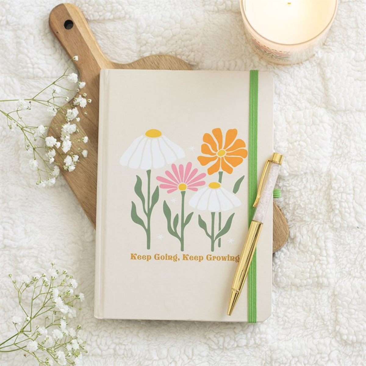 A journal decorated with illustrated flowers and the phrase Keep Going, Keep Growing rests on a wooden board. A rose quartz pen lies atop it, next to a candle and sprigs of baby's breath on a soft white textured blanket.
