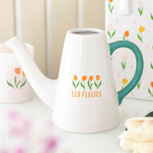White ceramic watering can with a green handle, ideal for tending plants, featuring four orange tulips above the words LES FLEURS. It rests on a white surface beside a candle and a floral-patterned object.