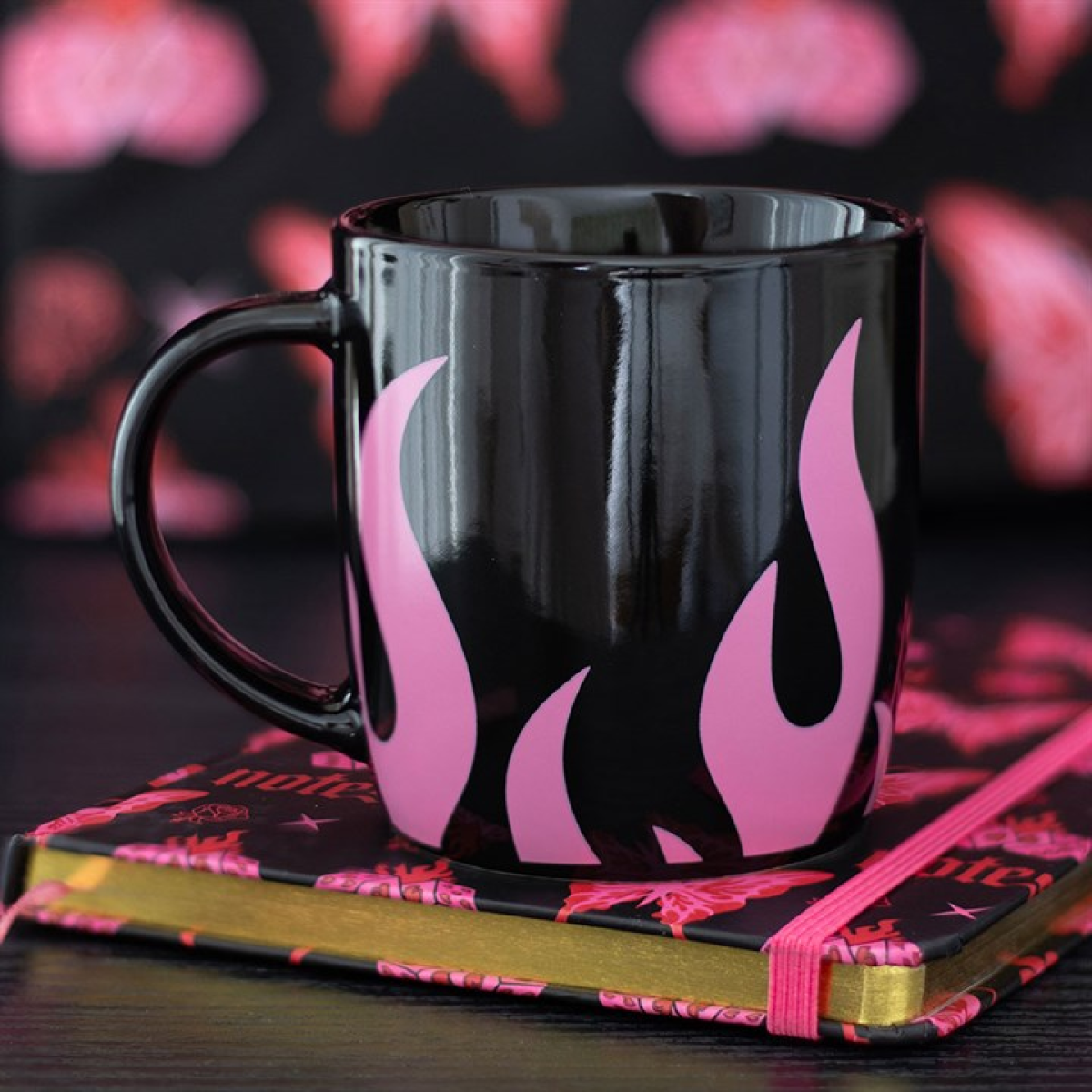 A striking coffee mug with glossy black ceramic and bright pink flames rests atop a closed notebook trimmed in gold and pink, set against a blurred backdrop of soft pink and black patterns.