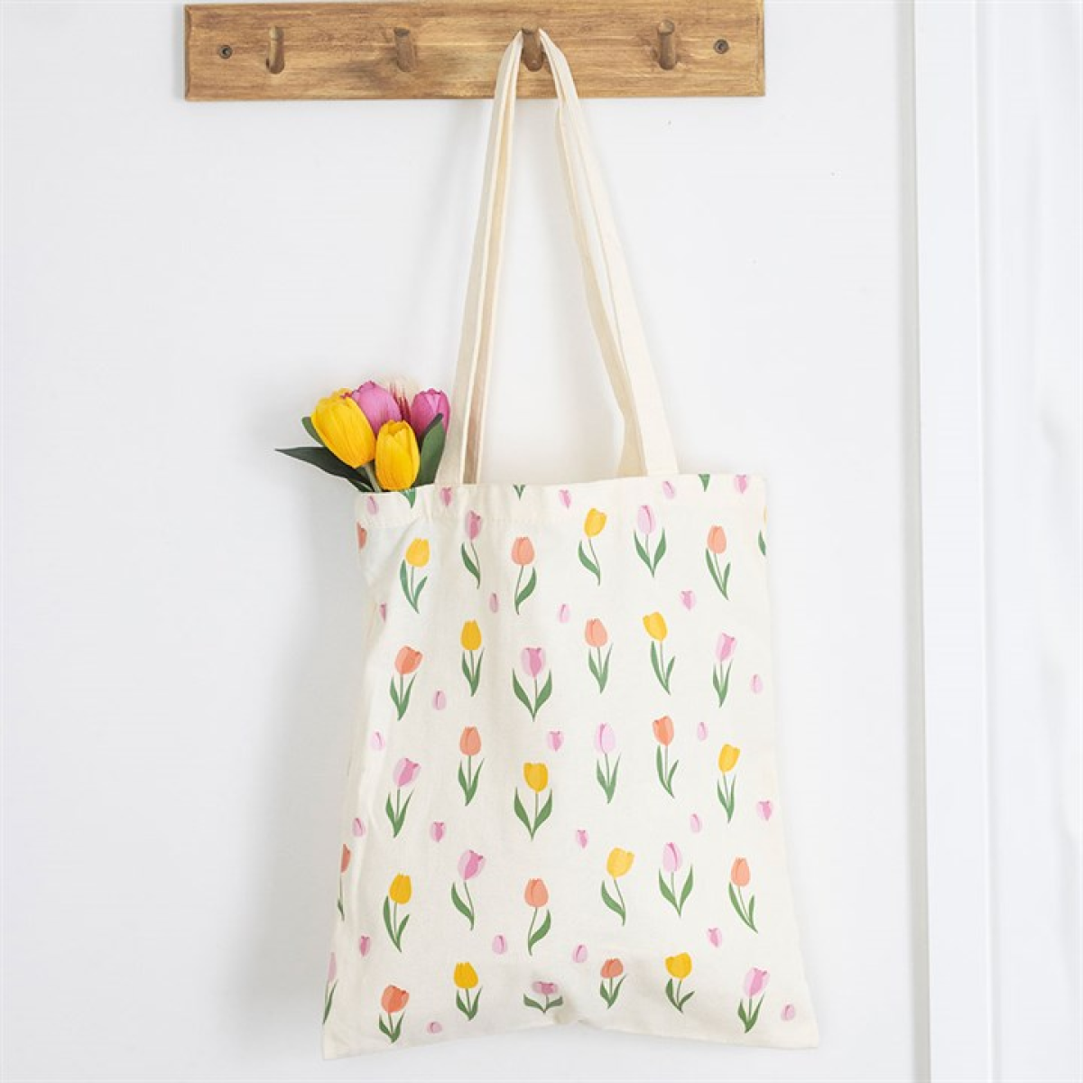 A cream-coloured reusable tote bag with a tulip print hangs from a wooden hook. Inside, a small bouquet of pink and yellow tulips with green leaves peeks out against a white wall background.