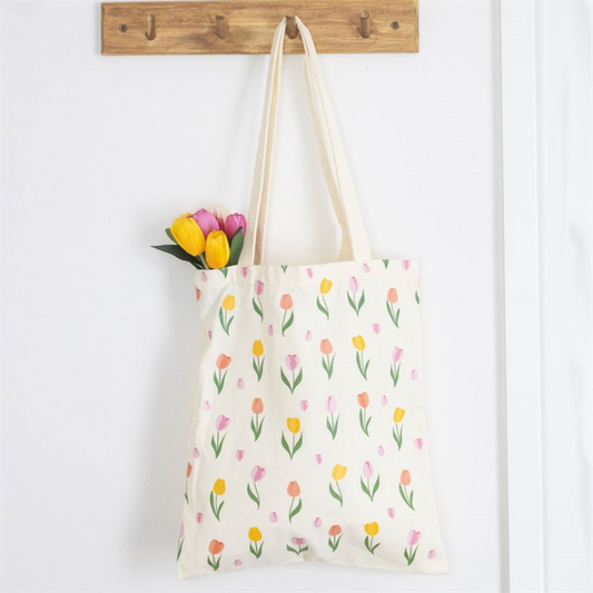 A cream-coloured reusable tote bag with a tulip print hangs from a wooden hook. Inside, a small bouquet of pink and yellow tulips with green leaves peeks out against a white wall background.