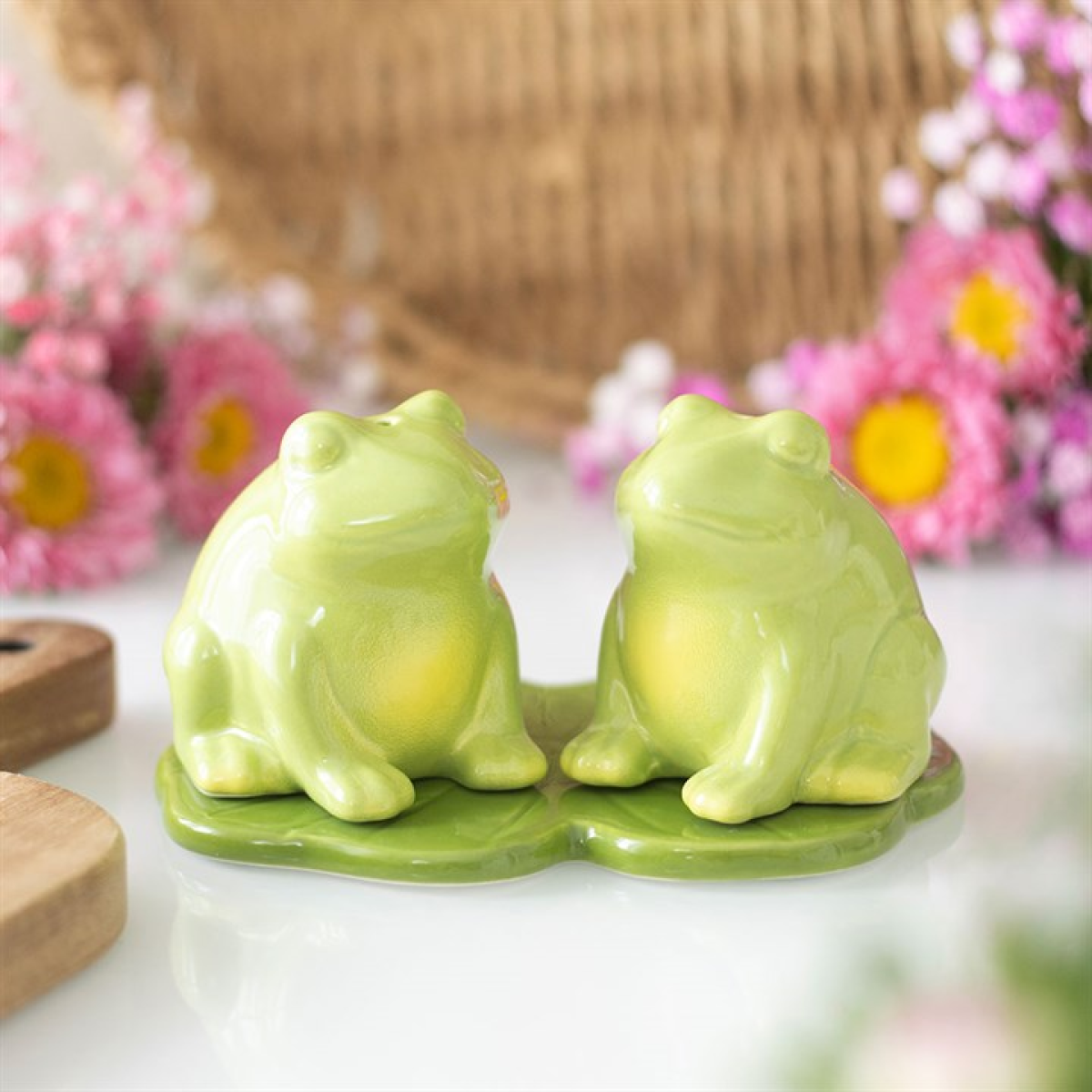 Two glossy ceramic frog salt and pepper shakers sit facing forward on a green base. Pink and yellow flowers blur in the background, adding cheerful charm to your kitchen décor.