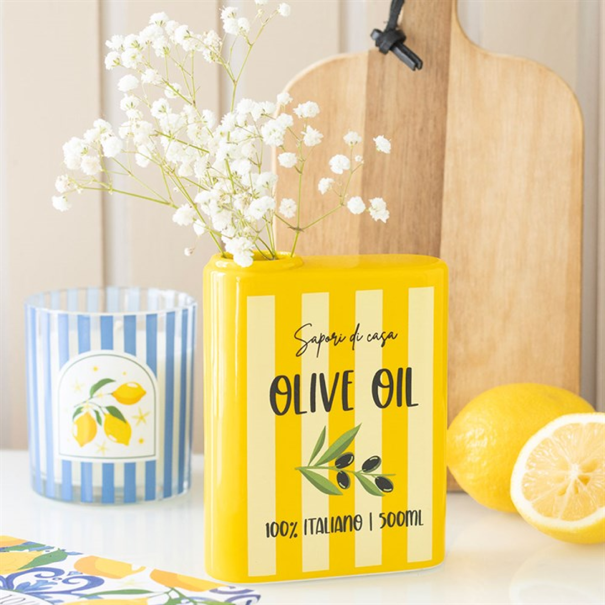 A yellow and white striped ceramic vase with a floral design—evoking Italian kitchen decor—holds white baby's breath. Close by are a sliced lemon, wooden board, and a blue glass decorated with a lemon motif.