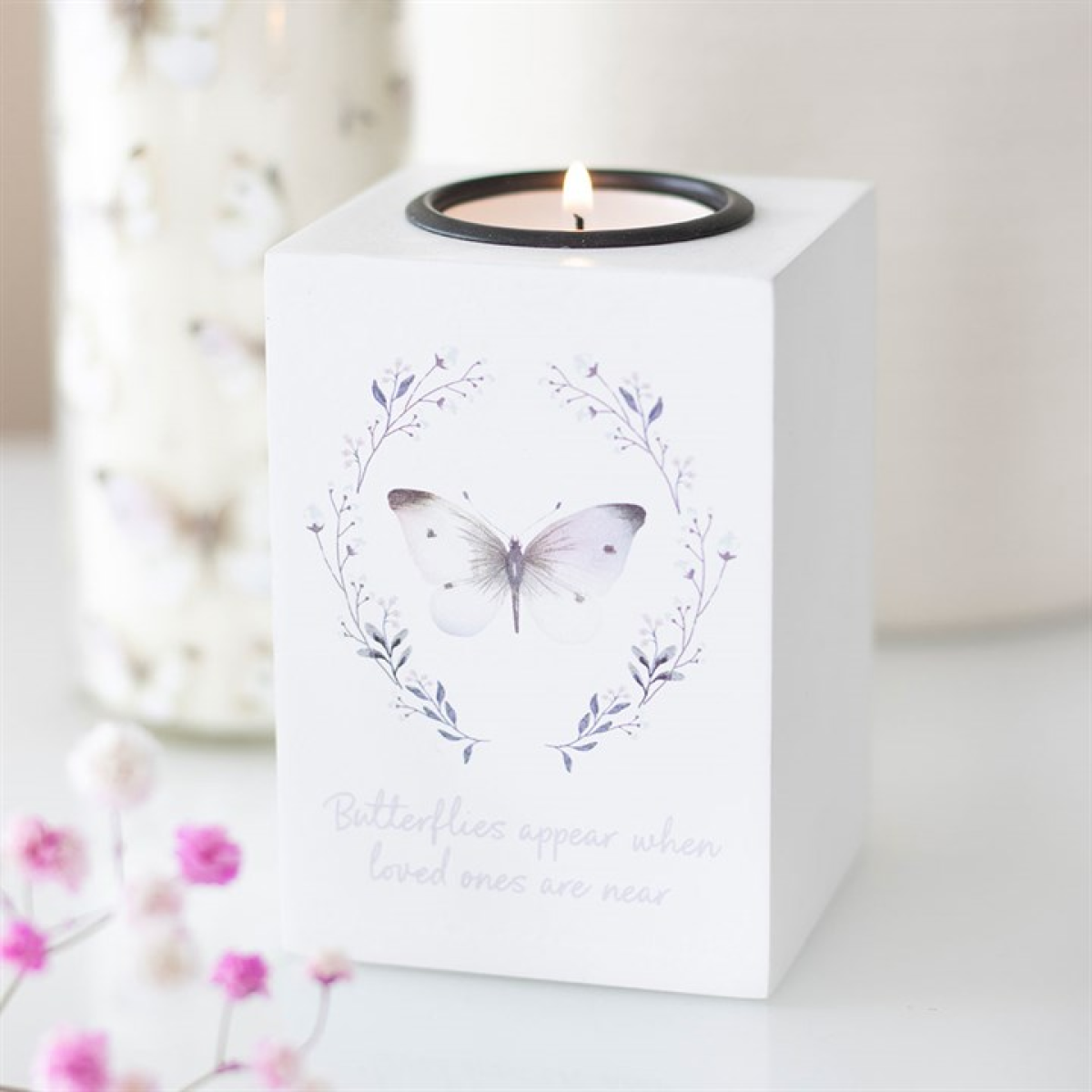 A white tealight holder with a lit candle, adorned with butterfly designs and a floral wreath. Beneath is the phrase “Butterflies appear when loved ones are near.” Nearby pink flowers make it an ideal remembrance gift.