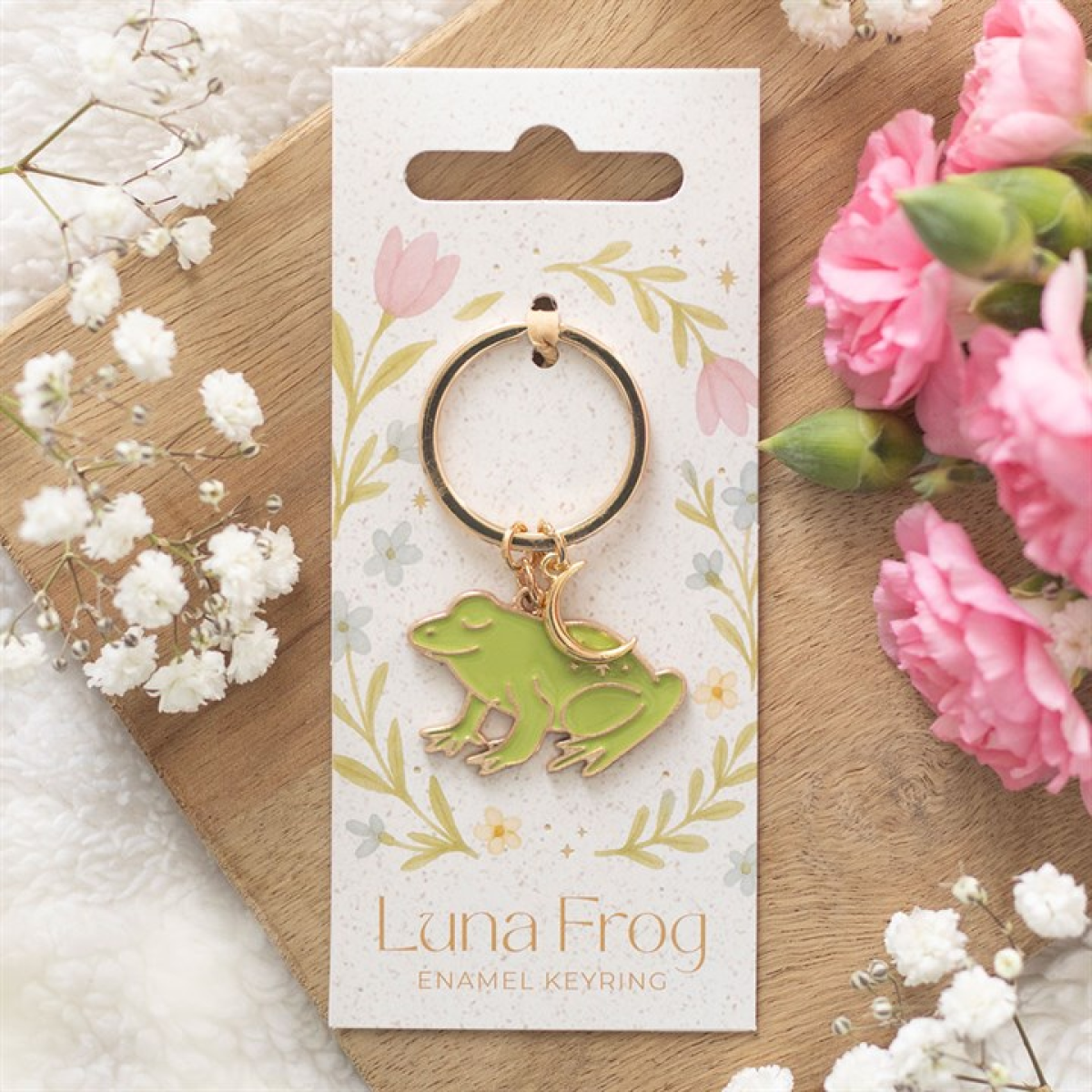 A gold-rimmed enamel frog keyring with a green charm sits on a floral card labelled “Luna Frog Enamel Keyring.” The card rests on wood, surrounded by pink flowers and white baby's breath.