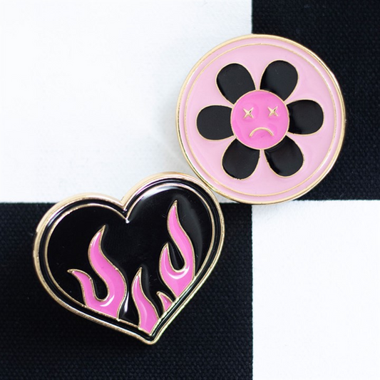 Two enamel pins on a black and white chequered background: a burning heart pin with black and pink flames, and a sad flower pin—a pink Y2K badge featuring a black flower with a sad face in the centre.