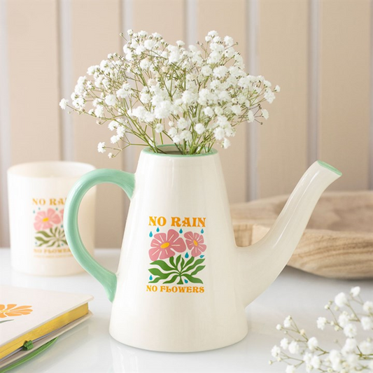 A cream-coloured ceramic watering can with a mint-green handle and spout holds white baby's breath flowers. Ideal for plant lovers, the can says NO RAIN NO FLOWERS above a pink and orange floral motif. Nearby: candle, book.