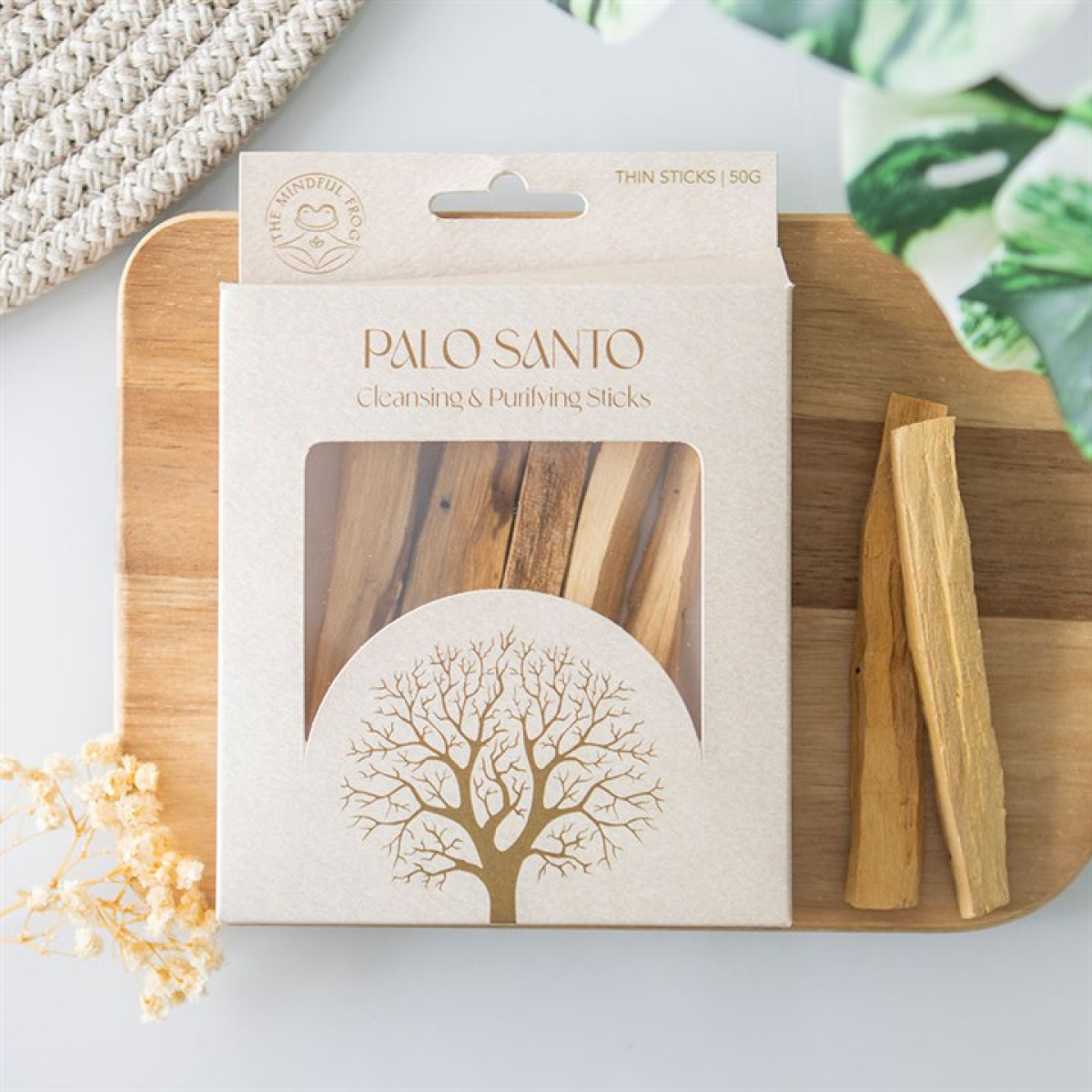 A box labelled Palo Santo Cleansing & Purifying Sticks rests on a wooden board. Two purifying palo santo sticks lie next to it, surrounded by green leaves, a woven mat, and dried flowers.