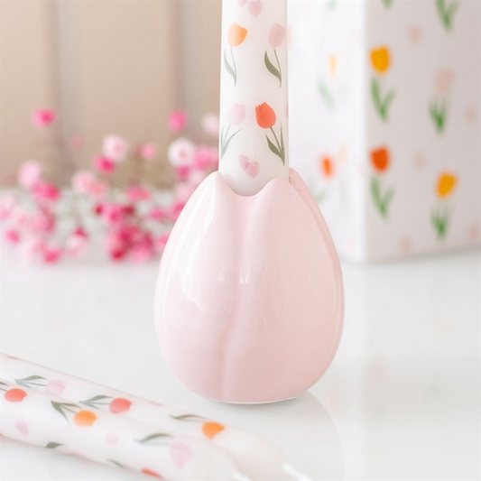 A pale pink ceramic candle holder, shaped like a tulip, holds a white candle decorated with pink, orange, and green florals. Subtle springtime touches such as blurred flowers and a matching floral box complete the background.