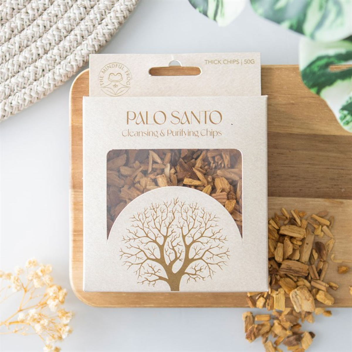 A box of Palo Santo chips for spiritual cleansing rests on a wooden board featuring a tree design. Loose chips, a woven mat, green leaves, and dried white flowers create this tranquil meditation setting.