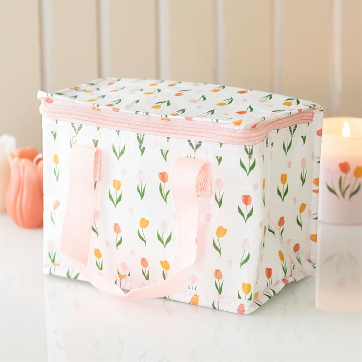 A rectangular insulated lunch bag with pink handles and a zip, adorned with a vibrant, unidentified floral pattern, rests on a white surface. In the background are a lit candle and an object shaped like a tulip.
