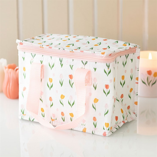 A rectangular insulated lunch bag with pink handles and a zip, adorned with a vibrant, unidentified floral pattern, rests on a white surface. In the background are a lit candle and an object shaped like a tulip.