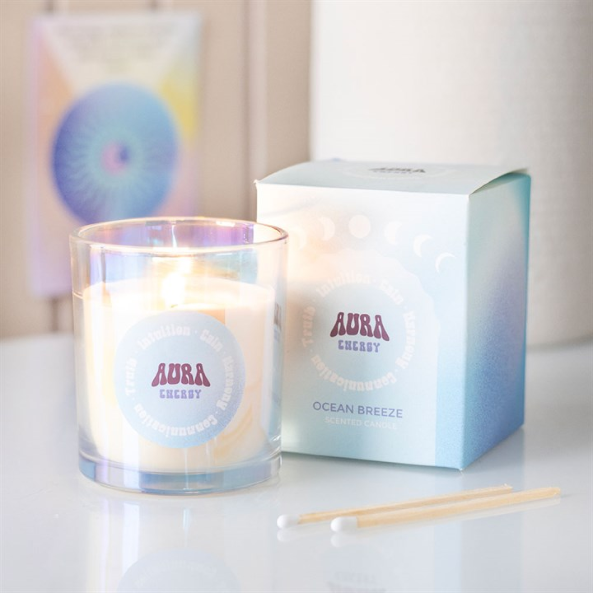 A lit “Aura Energy” Ocean Breeze scented candle in an iridescent glass jar sits beside its blue and white box on a glossy surface, with two wooden matches, a cotton bud, and a blurred card in the background.