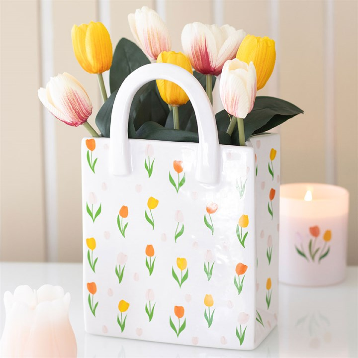 A quirky gift, this ceramic flower vase shaped like a shopping bag features a cheerful tulip print and holds artificial tulips in white, yellow, and pink. A lit candle and a white tulip-shaped ornament sit beside it.