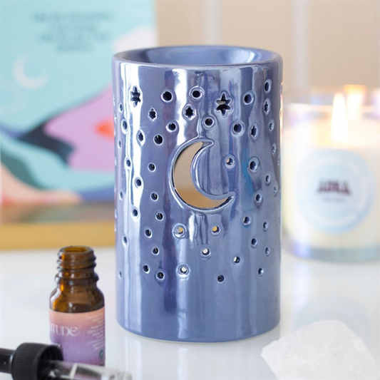 A glossy blue ceramic oil burner, ideal for fragrance oils or wax melts, features cut-out stars and a crescent moon. Nearby sit an essential oil bottle, a candle and crystal, set against a softly blurred, colourful background.