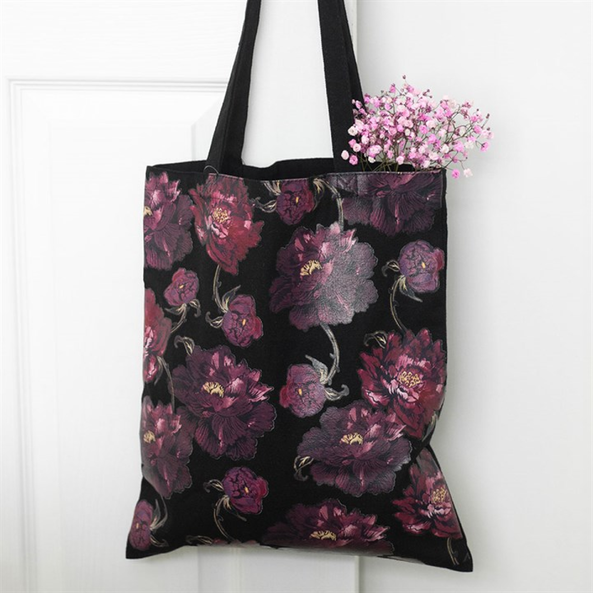 A black floral tote bag with dark purple and pink patterns hangs on a white door. Pink baby's breath flowers peek out, lending a delicate touch to the decorative accessory.