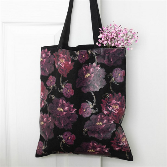A black floral tote bag with dark purple and pink patterns hangs on a white door. Pink baby's breath flowers peek out, lending a delicate touch to the decorative accessory.