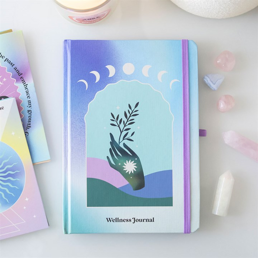 A pastel-coloured wellness journal featuring a hand holding a plant on the cover, surrounded by moon phases. Nearby are crystals, pink stones, a crystal chip pen, a candle and colourful cards on a white surface.