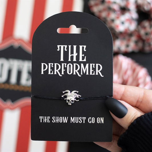 A hand with black matte nails holds a card labelled THE PERFORMER, adorned with a skull charm bracelet and a silver jester hat. The card says THE SHOW MUST GO ON. Red and white stripes blur in the background.