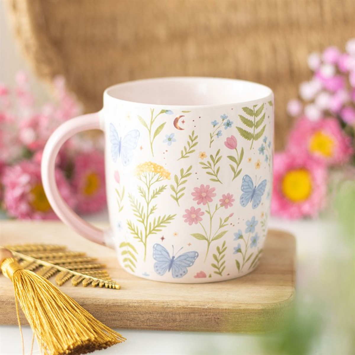 A pastel pink floral mug adorned with butterfly patterns rests on a wooden board. Pink and yellow flowers are blurred in the background; a golden tassel and wheat decoration sit beside this perfect tea lover’s gift.