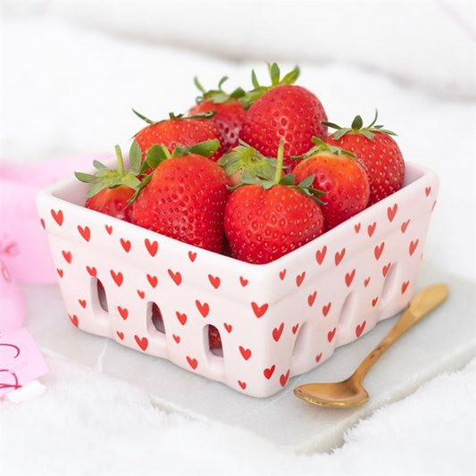 A square ceramic basket featuring a delightful heart print holds ripe strawberries with green stems. The dish rests on a light surface beside a small gold-coloured spoon, ideal for stylish storage or serving.