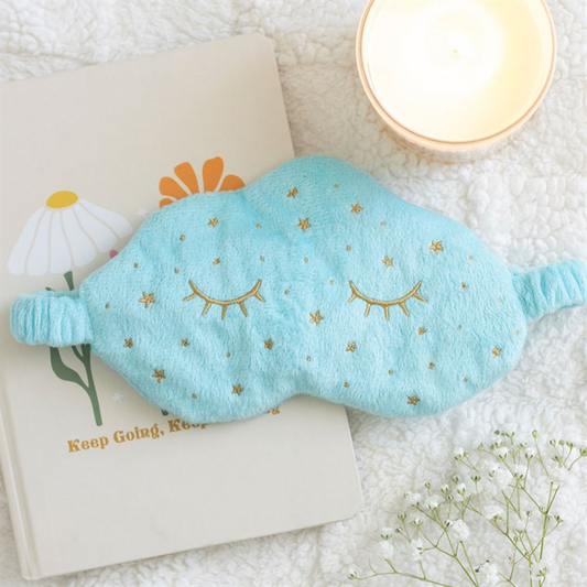 A soft blue sleep mask with gold embroidered stars and sleeping eyes lies on a notebook featuring a daisy and "Keep Going, Keep Growing" text—ideal for travel naps—next to a lit candle and small white flowers on a fluffy surface.