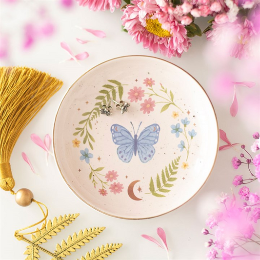 A round butterfly ceramic dish decorated with blue butterflies, pink and blue flowers, green ferns, and a crescent moon is surrounded by pink blossoms, gold fern leaves, and a tassel—a dreamy gift for nature lovers.
