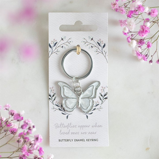 A sentimental gift, this silver butterfly keyring is attached to a floral card that reads, “Butterflies appear when loved ones are near,” bordered by pink and white flowers on a pale marble surface.