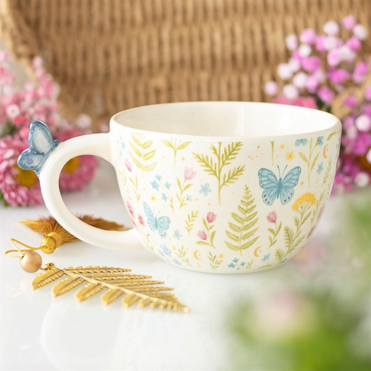 A white ceramic butterfly mug, decorated with blue butterflies and pastel flowers, rests on a white surface. Ideal for garden enthusiasts, its handle is topped with a blue butterfly. Pink flowers and a gold leaf ornament lie nearby.