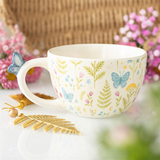 A white ceramic butterfly mug, decorated with blue butterflies and pastel flowers, rests on a white surface. Ideal for garden enthusiasts, its handle is topped with a blue butterfly. Pink flowers and a gold leaf ornament lie nearby.