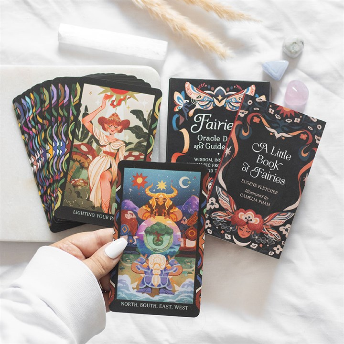 A hand holds a colourful fairy-themed tarot card. Other divination cards, a fairy oracle deck box, a Little Book of Folklore Fairies, and pastel crystals rest on a white surface in bright, soft lighting.