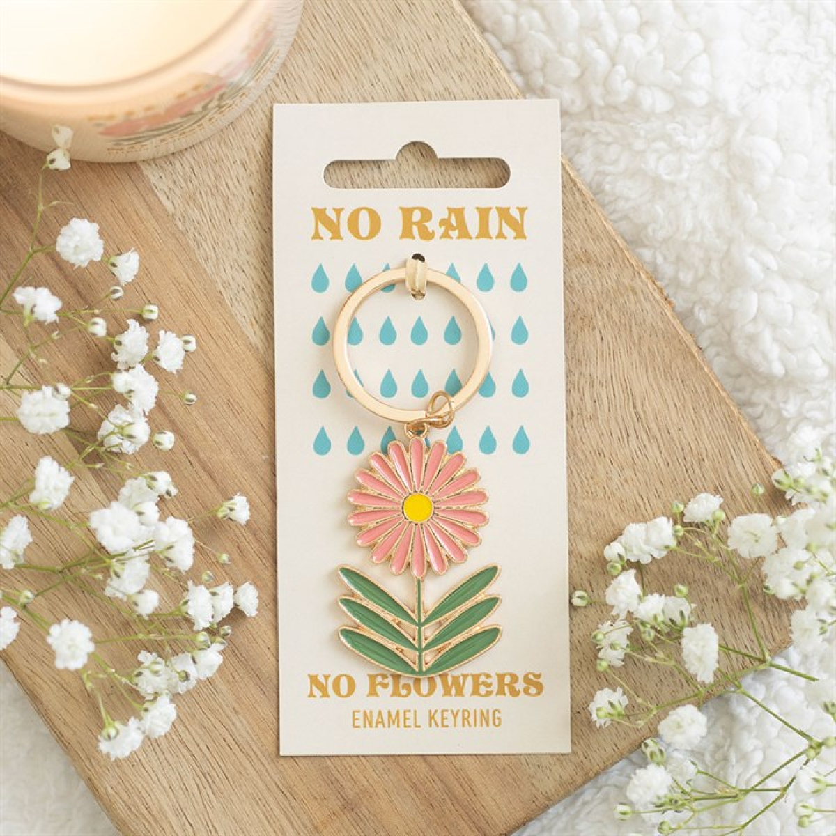 A charming keyring featuring a pink and yellow daisy in a retro enamel style is fastened to a card with blue raindrops and the message “NO RAIN NO FLOWERS.” White baby's breath surrounds it on a wooden surface.