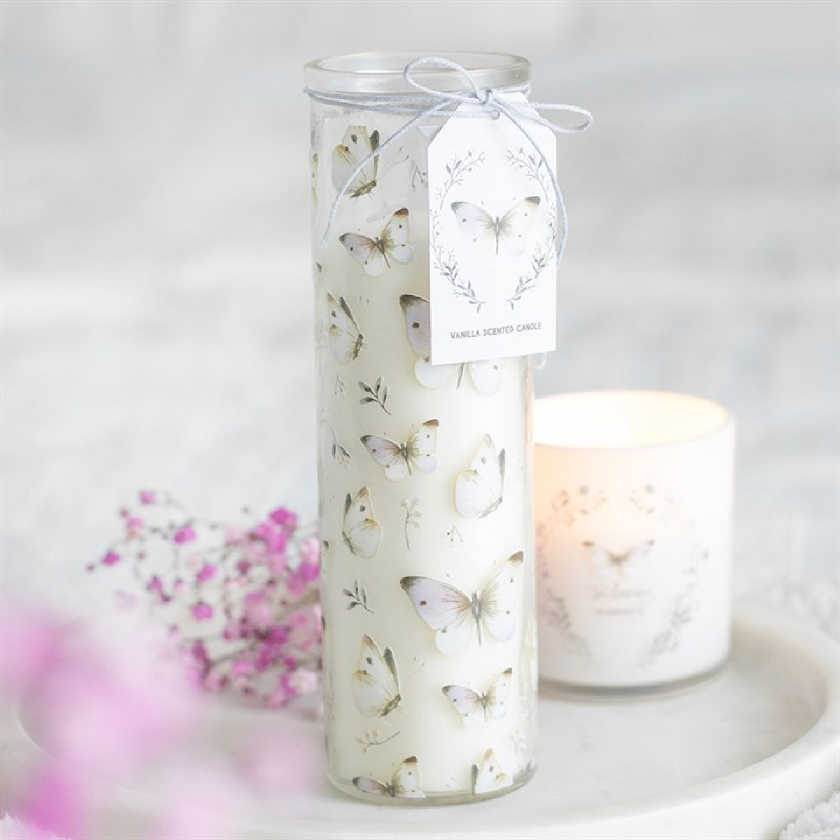 A tall, white vanilla-scented candle featuring a delicate butterfly design and a tag tied with silver string stands on a white surface. Pink baby's breath and a lit candle in frosted glass adorn the background.