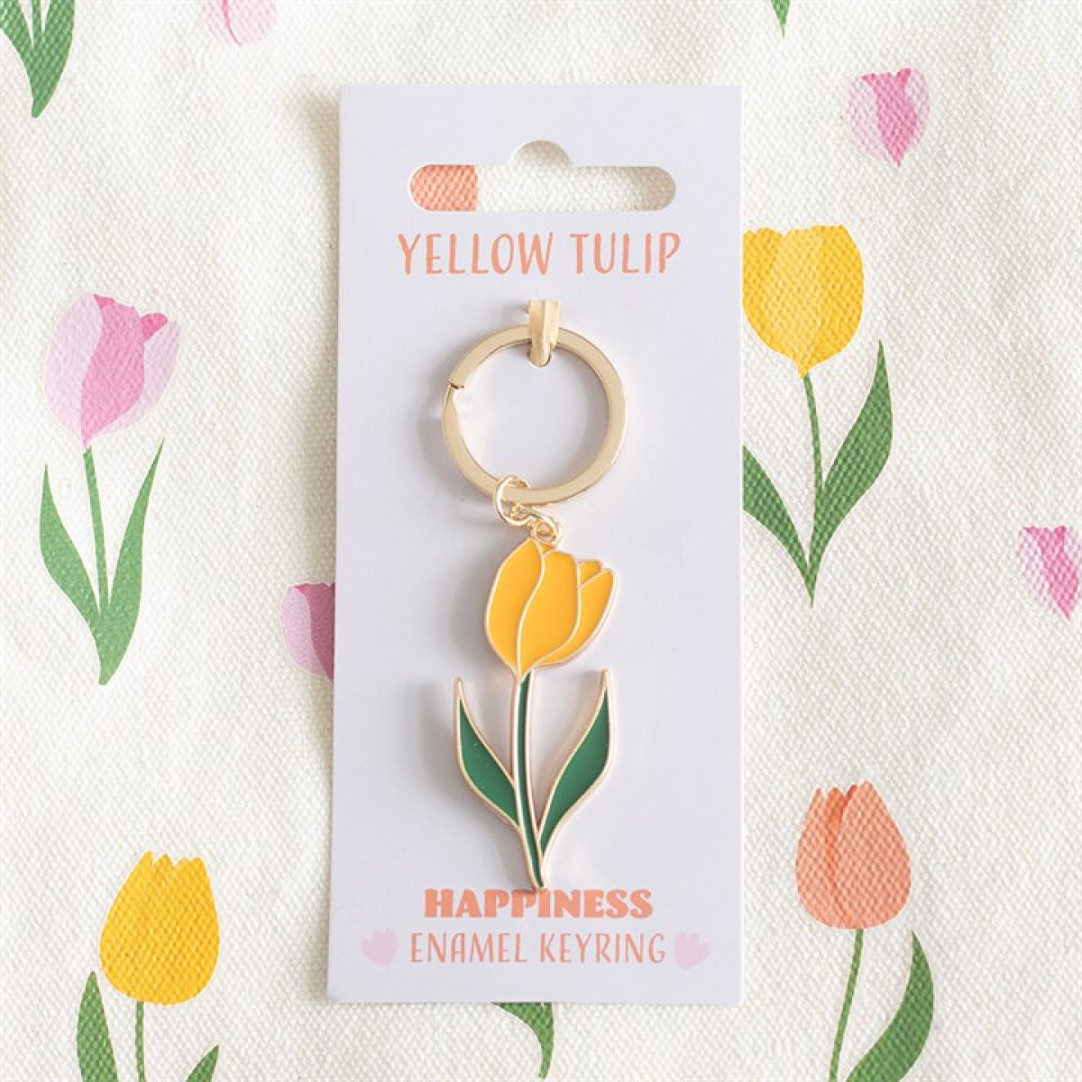 A golden-yellow tulip keyring with a delightful enamel charm is affixed to a card reading YELLOW TULIP and HAPPINESS ENAMEL KEYRING, making an adorable gift. The background features a vibrant floral tulip pattern.