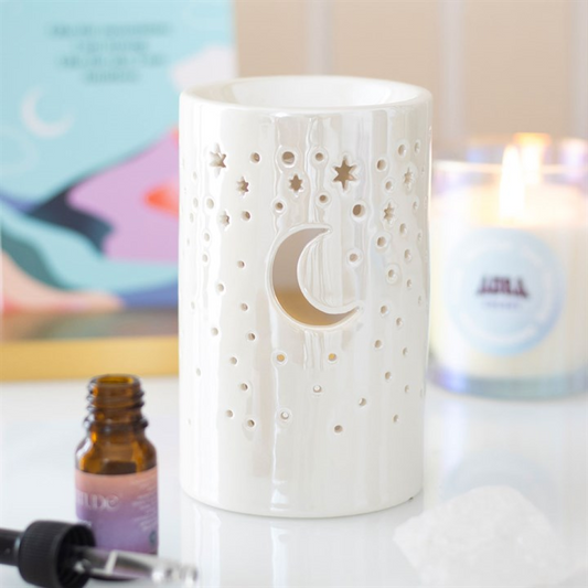 A white ceramic burner with star and moon cut-outs sits on a table next to a lit candle, fragrance oils in an amber dropper bottle, and a clear crystal. The softly blurred background lends a calming feel.