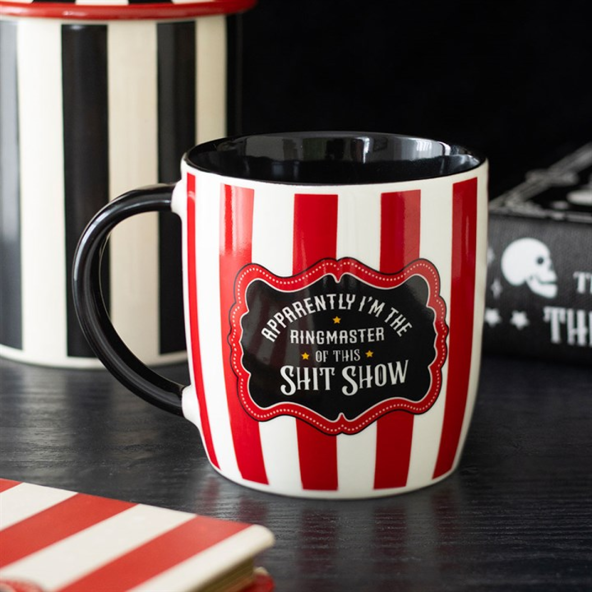 A humorous coffee mug with vertical red and white stripes has a black handle and a label reading, “Apparently I’m the ringmaster of this shit show.” Other circus-themed mugs and skull items are blurred in the background.