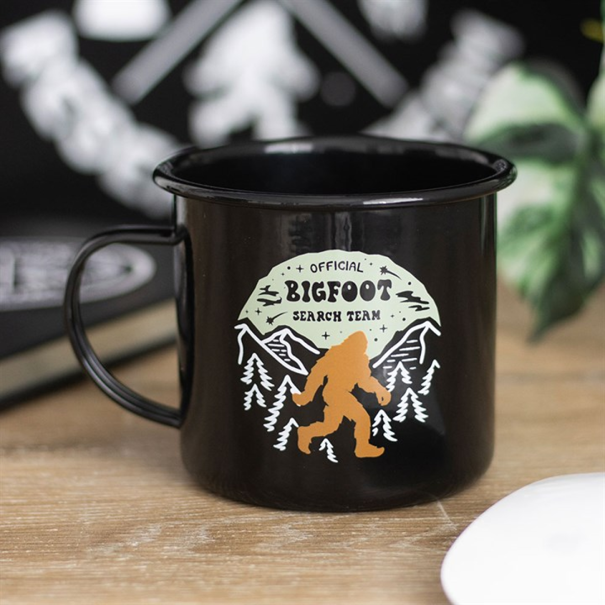 A black enamel-style mug features a mountain scene with pine trees, a striking orange Bigfoot silhouette, and the phrase “Official Bigfoot Search Team”—an ideal companion for your next cryptid stakeout.