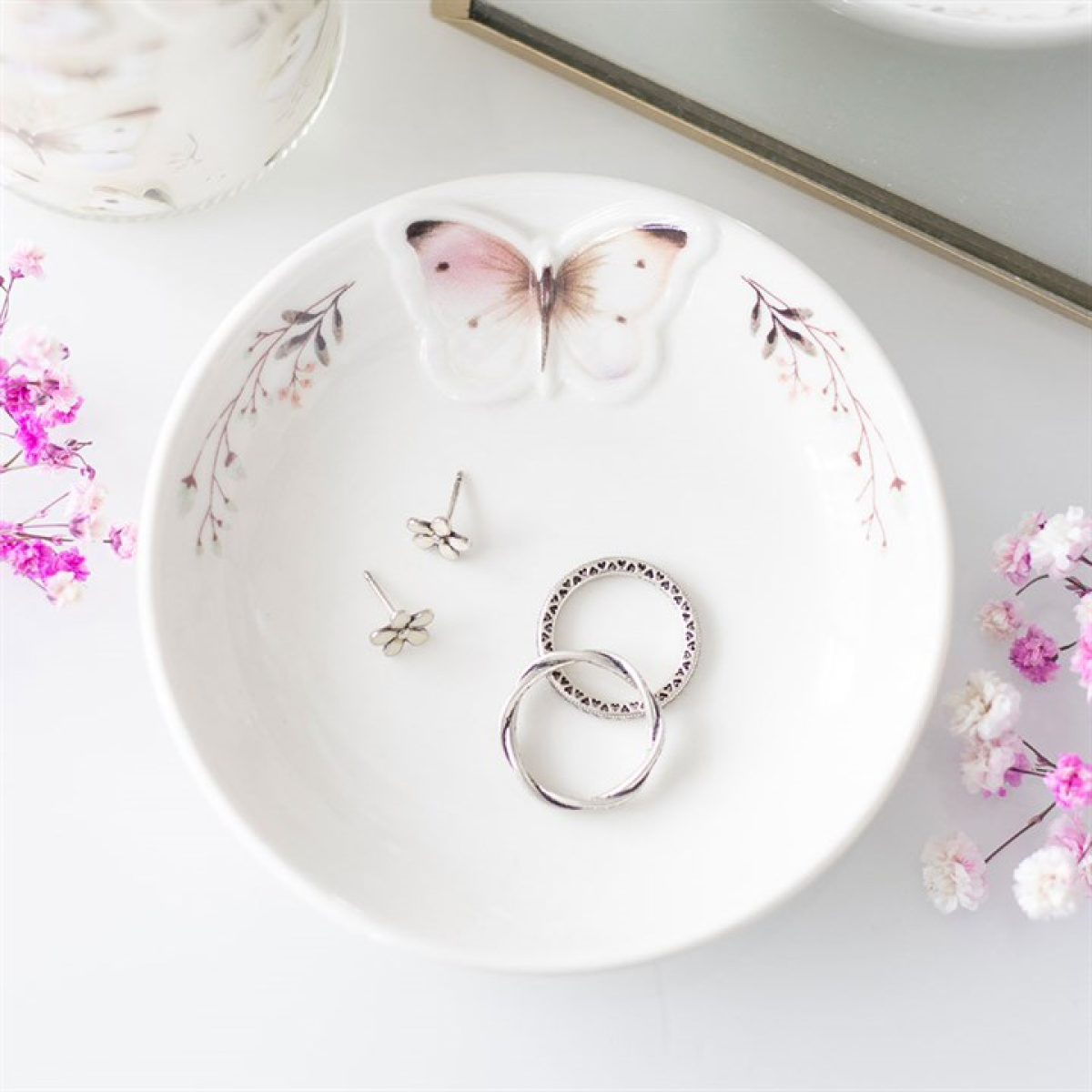 A ceramic trinket dish featuring a painted butterfly and pink flowers holds silver rings and flower-shaped studs. Ideal as bedside table decor, it is styled with pink and white dried flowers on a white surface.