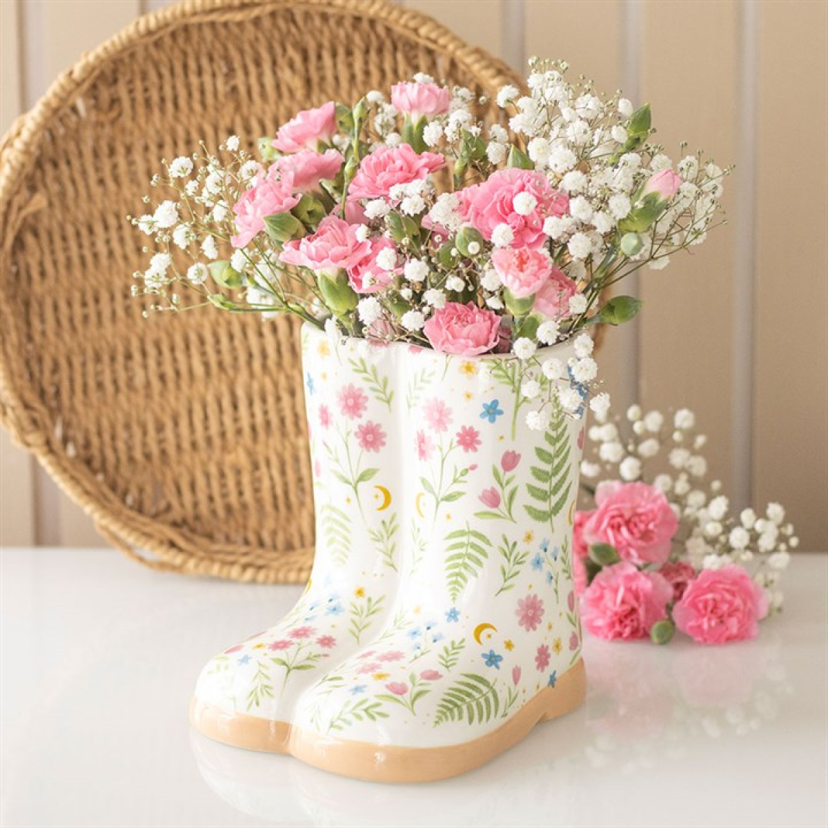 A ceramic welly boot planter decorated with a floral pattern holds pink carnations and white baby’s breath; additional flowers rest beside it, all set against a wicker backdrop and a beige panelled wall.
