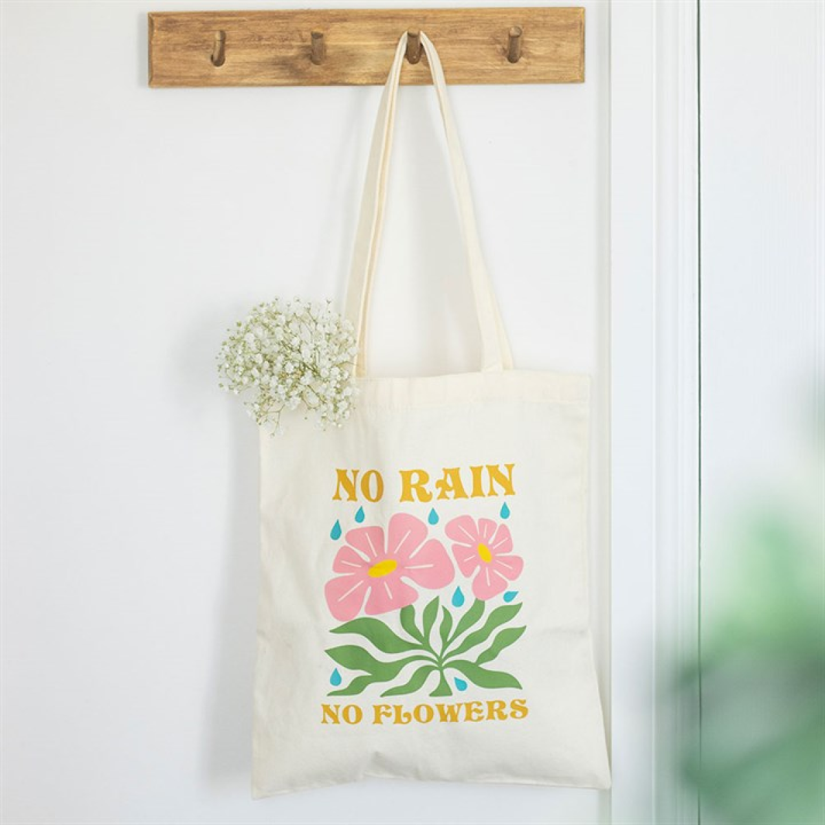 A stylish tote bag hangs on a wooden hook rack, adorned with pink flowers, blue raindrops, and "No Rain No Flowers" in yellow. Baby's breath flowers peep out of this lovely tote.